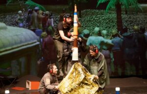 Reconstitution du lancement de la fusée Apollo 11 depuis Cap Canaveral avec assemblage de bouteilles thermos, lampe torche et couverture de survie sur fond de projection sur grand écran d'une foule constituée de petits personnages en plastique.