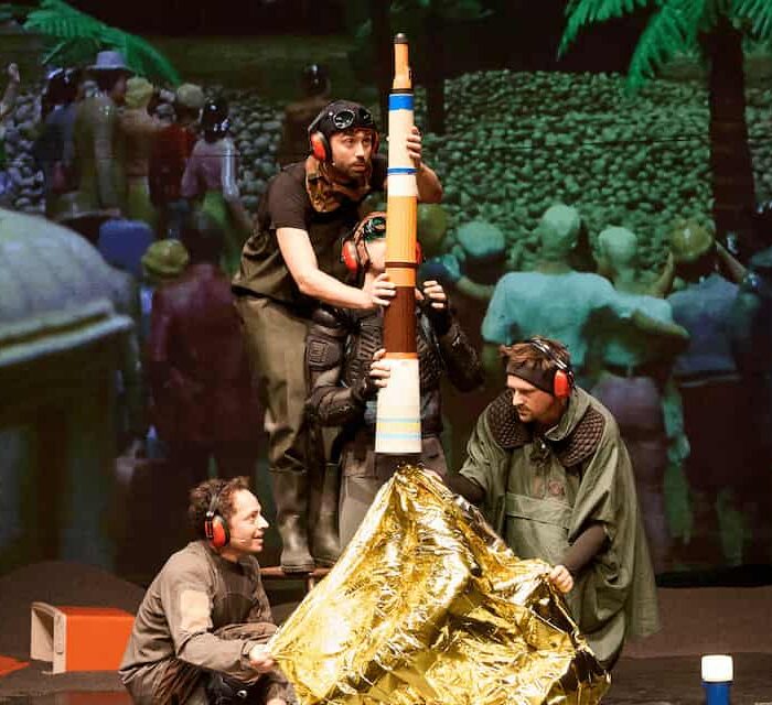 Reconstitution du lancement de la fusée Apollo 11 depuis Cap Canaveral avec assemblage de bouteilles thermos, lampe torche et couverture de survie sur fond de projection sur grand écran d'une foule constituée de petits personnages en plastique.