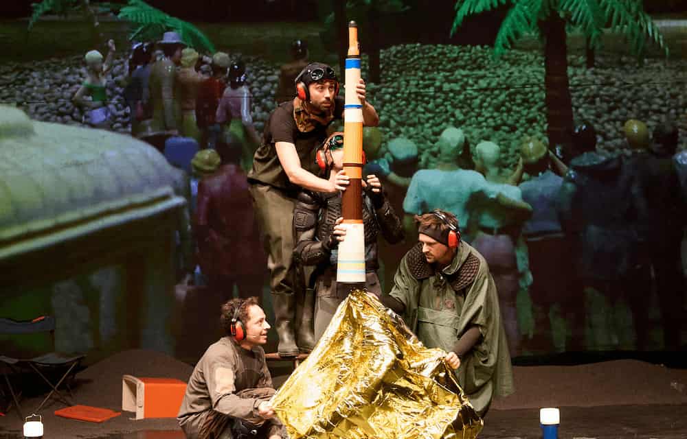 Reconstitution du lancement de la fusée Apollo 11 depuis Cap Canaveral avec assemblage de bouteilles thermos, lampe torche et couverture de survie sur fond de projection sur grand écran d'une foule constituée de petits personnages en plastique.