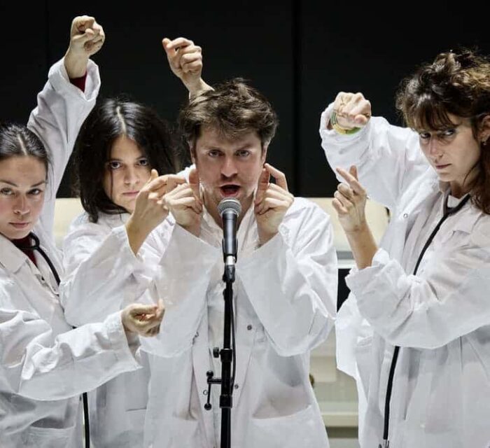 Un homme entouré de trois femmes chante dans un micro. Ils sont tous en blouse blanche et ont les bras en l'air. Ils claquent des doigts pour pulser le rythme.