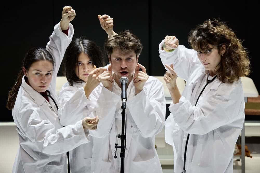 Un homme entouré de trois femmes chante dans un micro. Ils sont tous en blouse blanche et ont les bras en l'air. Ils claquent des doigts pour pulser le rythme.