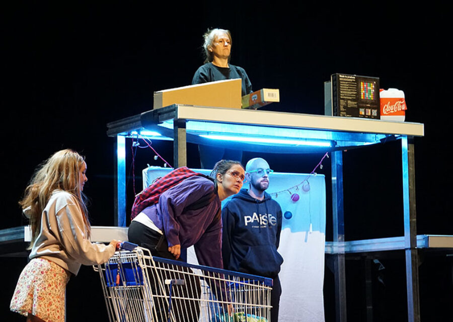 Quatre personnages dans un décor constitué de tables plus ou moins hautes symbolisant une caisse de supermarché. Une jeune femme dans un caddie dépose ses achats devant la caissière.